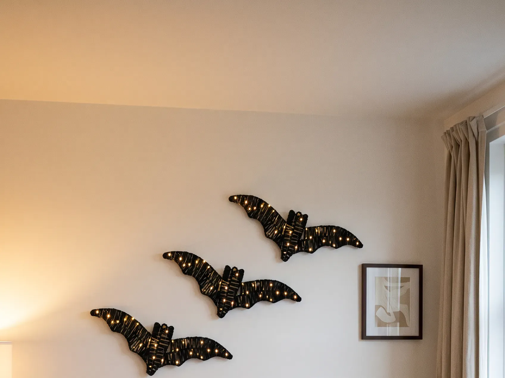 Three lit rattan bats arranged in a rising diagonal on a plain painted wall above a sofa, no-nail indoor Halloween decorations for a rental