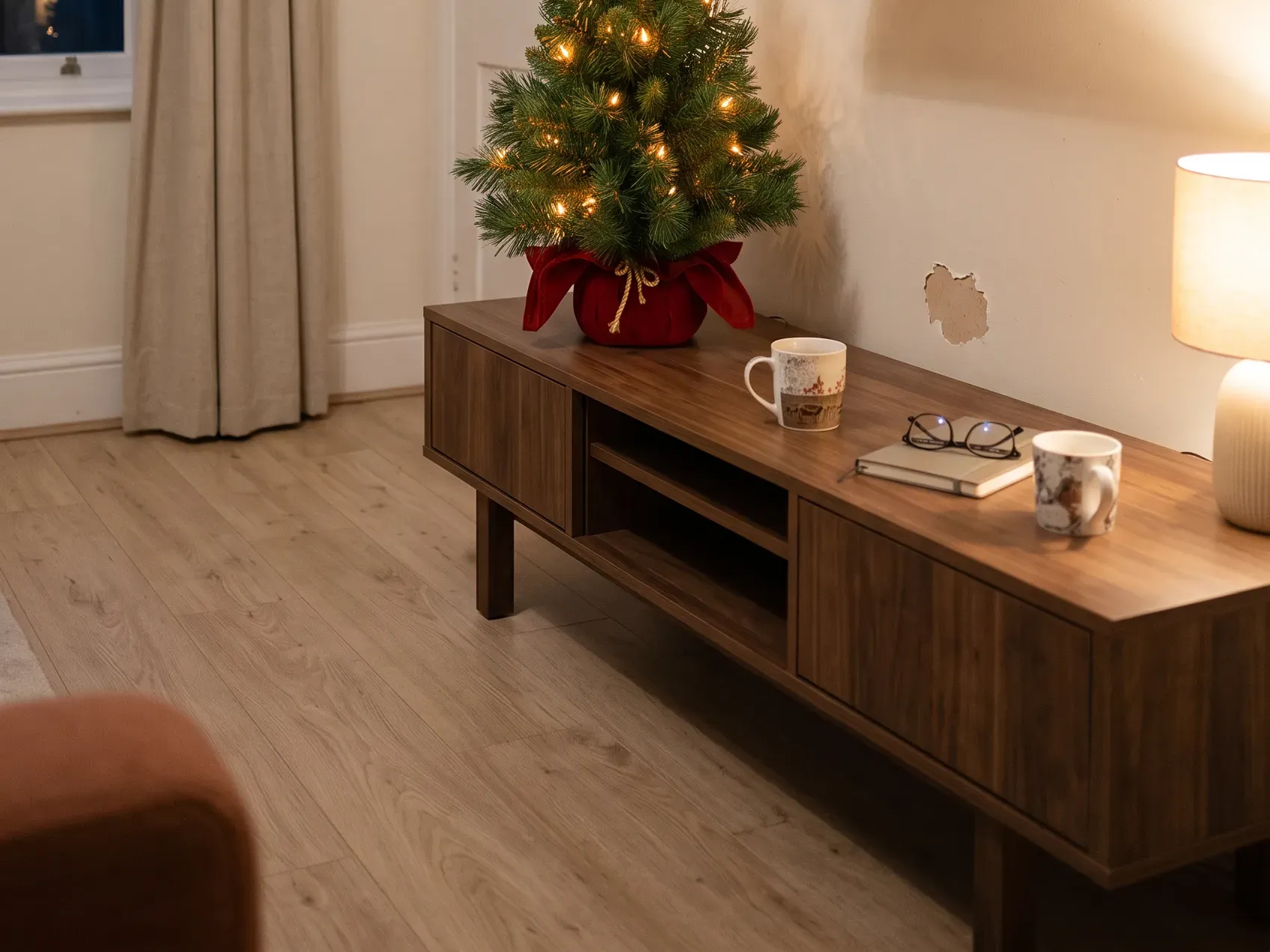 Small pre-lit tabletop Christmas tree on a console table beside a stack of books, with a set of small bottle brush trees on the shelf below, small apartment Christmas decor for a room with no floor space